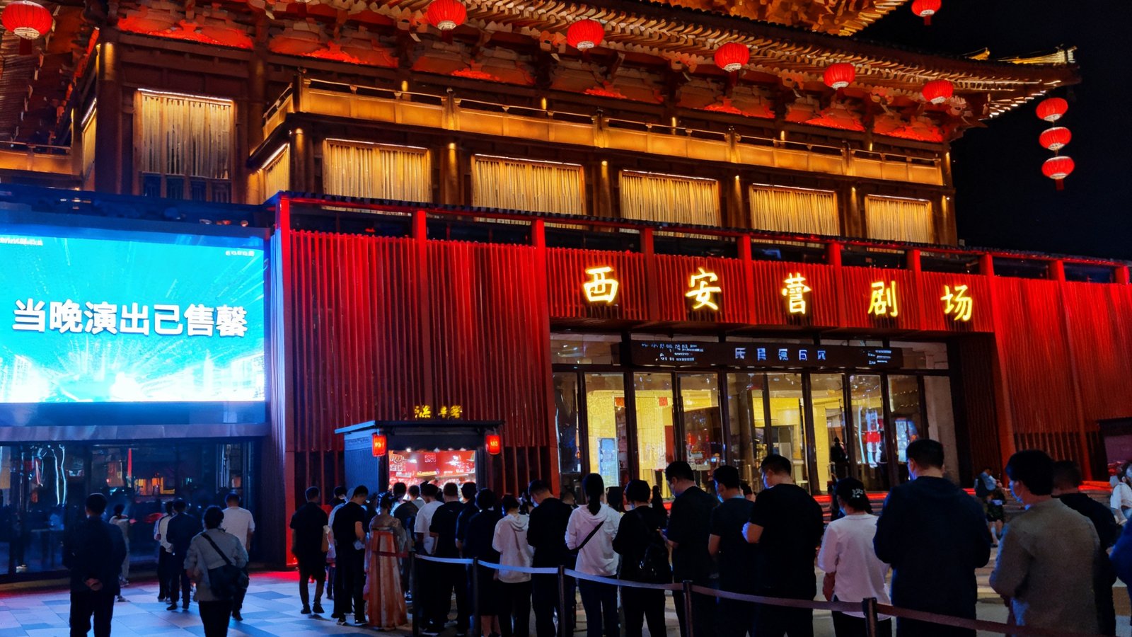西安大唐不夜城全息剧场外观夜景，仿唐建筑门头悬挂红色灯笼，入口处排起长队，LED屏显示当晚演出已售罄
